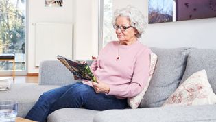 Ältere Dame mit Hausnotruf-Knopf an einer Kette liest auf der Couch eine Zeitschrift.