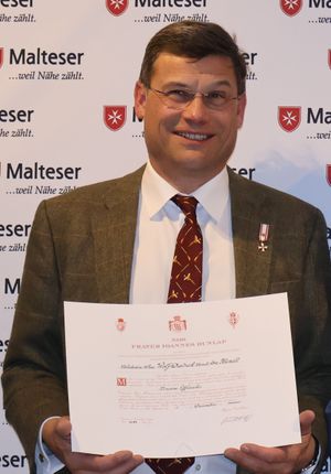 Herr mit roter Krawatte und großer Urkunde steht vor einer Wand mit vielen Malteser Logos