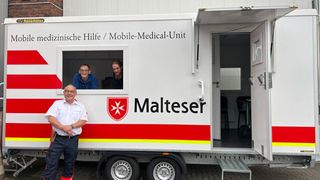 Anhänger der Mobilen Malteser Hilfer wo drei Helfer davor zu sehen sind
