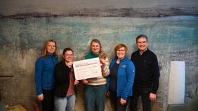 Fünf Personen stehen in einem Raum vor einer blau-grau strukturierten Wand und halten gemeinsam einen großen Spendenscheck. Zwei der Personen tragen blaue Jacken mit Malteser-Aufdruck. Auf dem Scheck steht „Spendenübergabe der Weihnachtsaktion 2023“ sowie die übergebene Summe und Unterschriften. Die Gruppe posiert für ein gemeinsames Foto der Spendenübergabe.