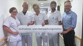 Fünf Klinikmitarbeitende stehen im Flur und halten ein Schild mit der Aufschrift „Aufnahme- und Überwachungsstation“.