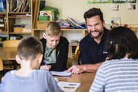 Mehrere Kinder sitzen gemeinsam an einem Tisch in einem Klassenraum, während ein lächelnder Erwachsener sie bei einer Schreibaufgabe unterstützt. Im Hintergrund sind Regale mit Büchern und Unterrichtsmaterialien zu sehen.