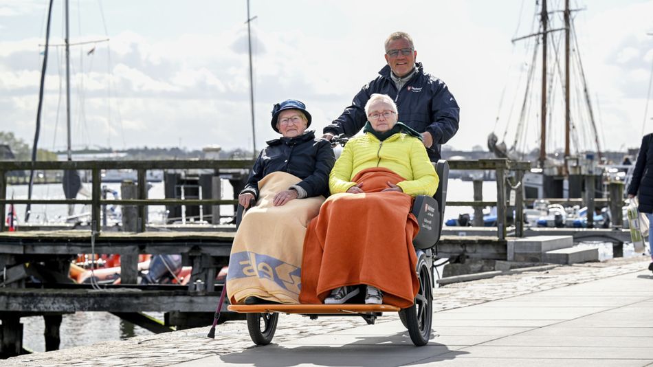 Zwei ältere Frauen sitzen nebeneinander in einem dreirädrigen Rollstuhl-Fahrrad und sind mit Decken zugedeckt, während ein Mann sie von hinten schiebt. Im Hintergrund ist ein Hafen mit Segelbooten und Stegen zu sehen, und die Gruppe bewegt sich entlang einer gepflasterten Uferpromenade.