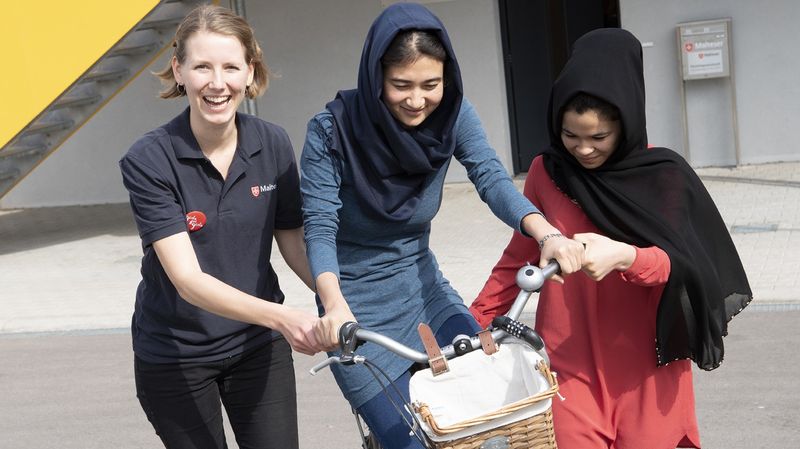 Eine lächelnde Frau bringt zwei jungen geflüchteten Frauen das Fahrradfahren bei. 