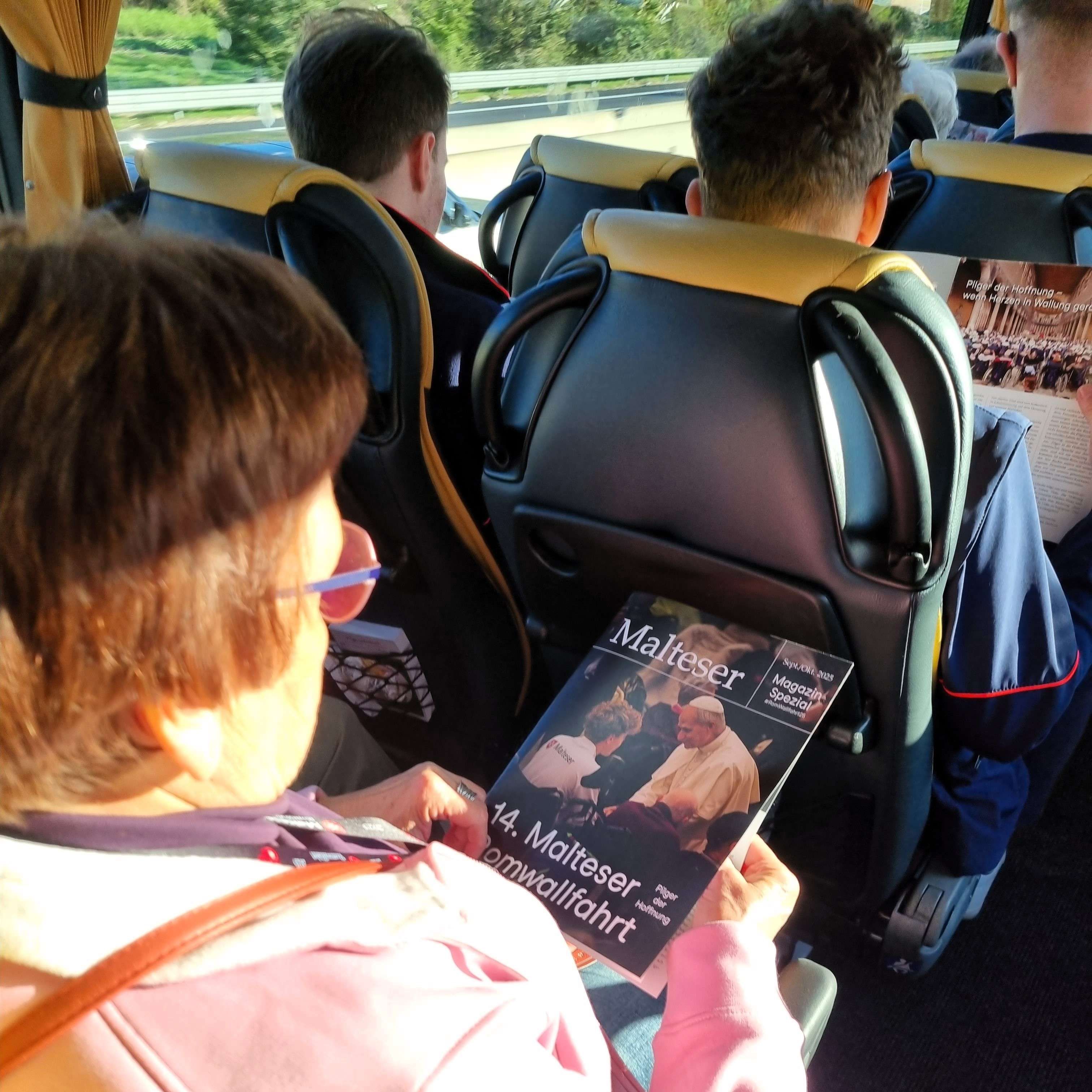 Person mit Brille liest Magazin zur Romwallfahrt im Bus, andere Fahrgäste sitzen vor ihr, Busfenster mit Landschaft im Hintergrund.