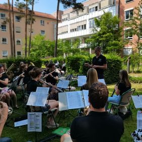 Blasorchester spielt im grünen Innenhof eines Klinikgeländes vor einem Dirigenten im schwarzen T-Shirt.