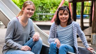 Eine junge Schulbegleiterin sitzt mit einem Mädchen auf dem Spielplatz.