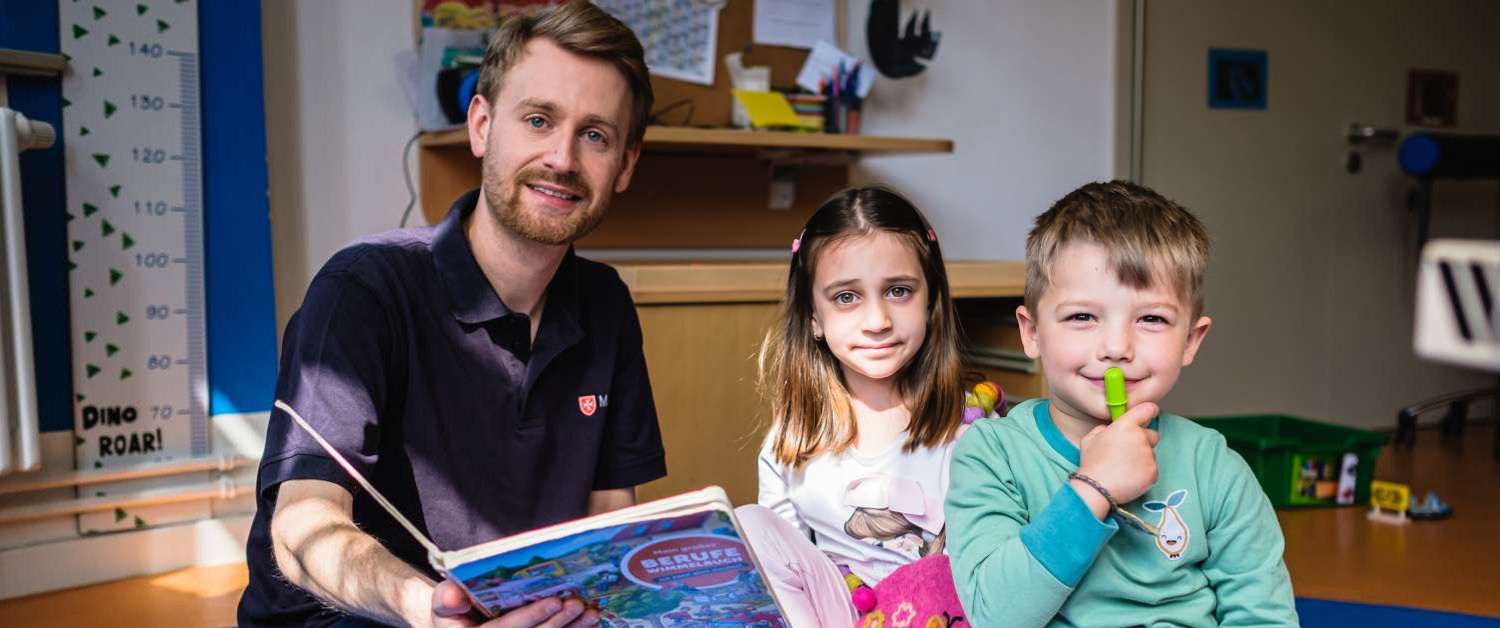 Ein Mann sitzt mit einem geöffneten Kinderbuch und zwei Kindern auf dem Boden