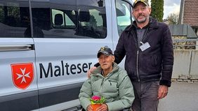 Ein Mann im Rollstuhl mit einem Blumentopf in der Hand, dahinter ein Mann in Malteserkleideung, dahinter ein Behindertentransportfahrzeug der Malteser.