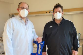 Arzt in weißem Kittel steht mit maskiertem Patienten im Patientenzimmer, beide halten gemeinsam Gehhilfen in der Hand.