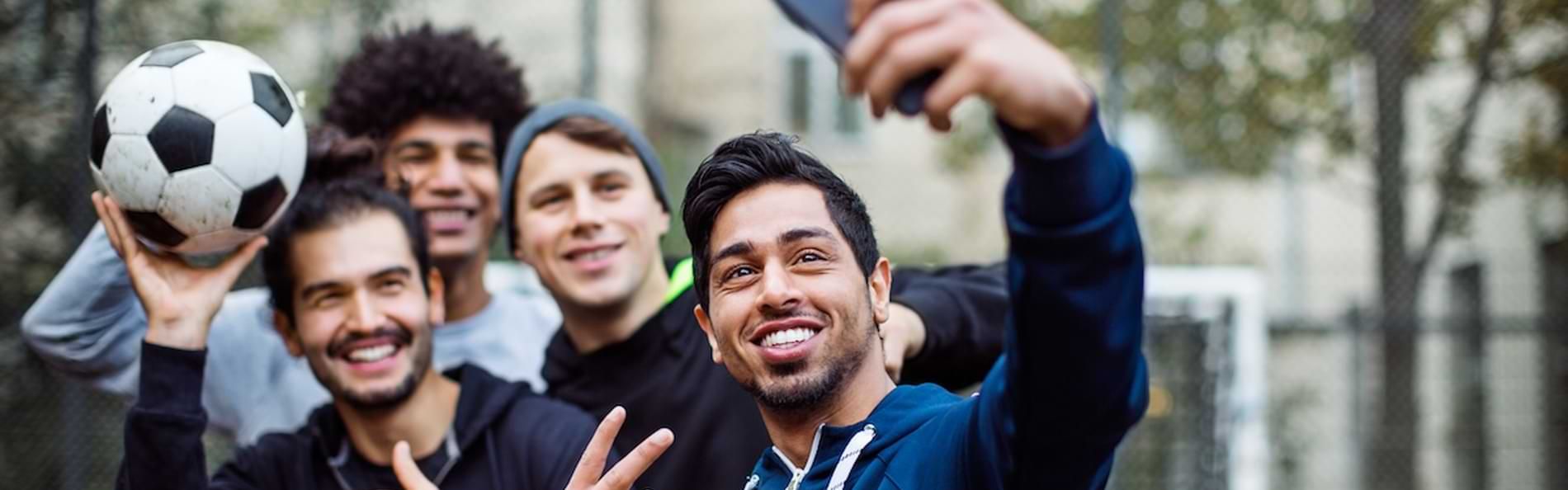 Eine Gruppe junger Männer unterschiedlicher Herkunft steht auf einem Bolzplatz und macht ein gemeinsames Selfie.