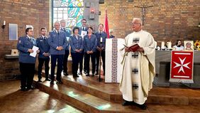 Ein Pfarrer steht im Vordergrund. Im Hintergrund stehen Menschen in Malteser-Kleidung.