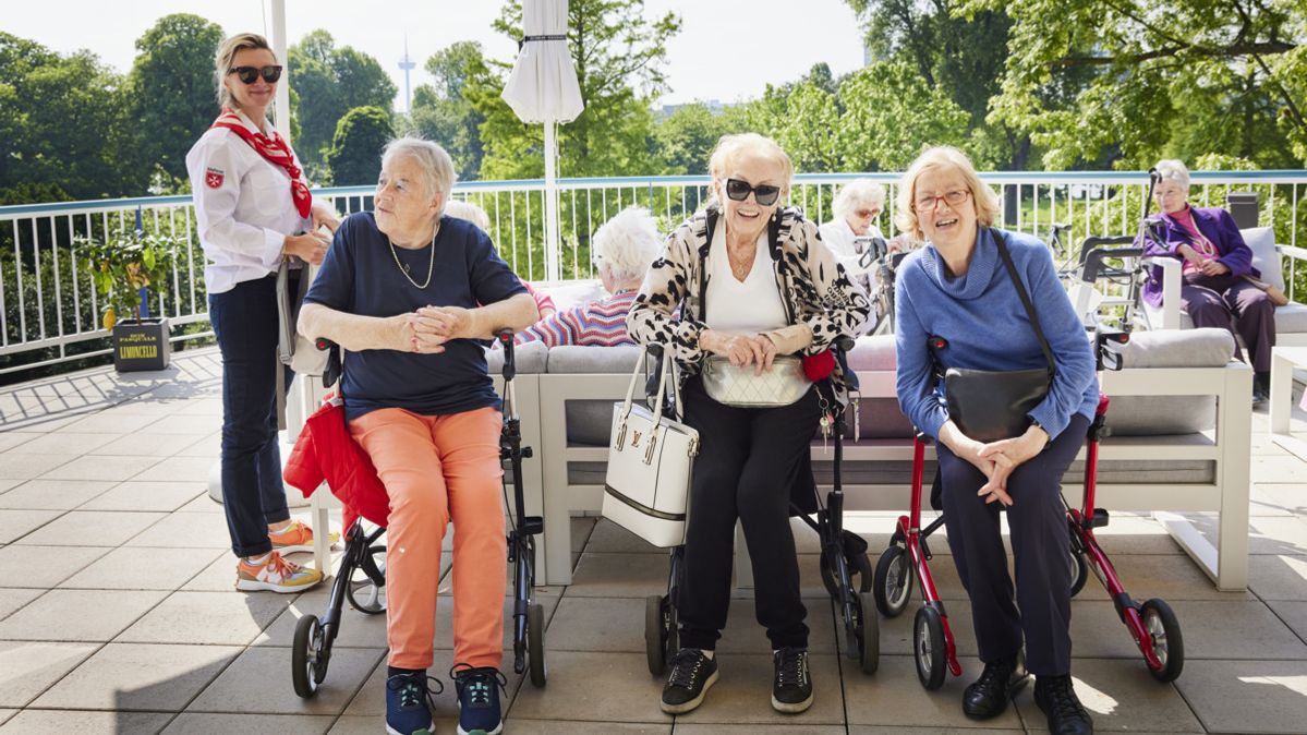Mehrere ältere Menschen sitzen und stehen gemeinsam auf einer Terrasse im Freien, einige nutzen Rollatoren oder Rollstühle. Eine Begleitperson steht daneben. Im Hintergrund sind Bäume und weitere Sitzgelegenheiten zu sehen.