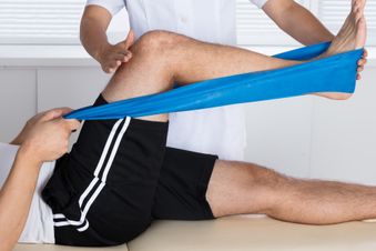 Physiotherapeutin unterstützt liegenden Patienten bei Knieübung mit blauem Fitnessband.