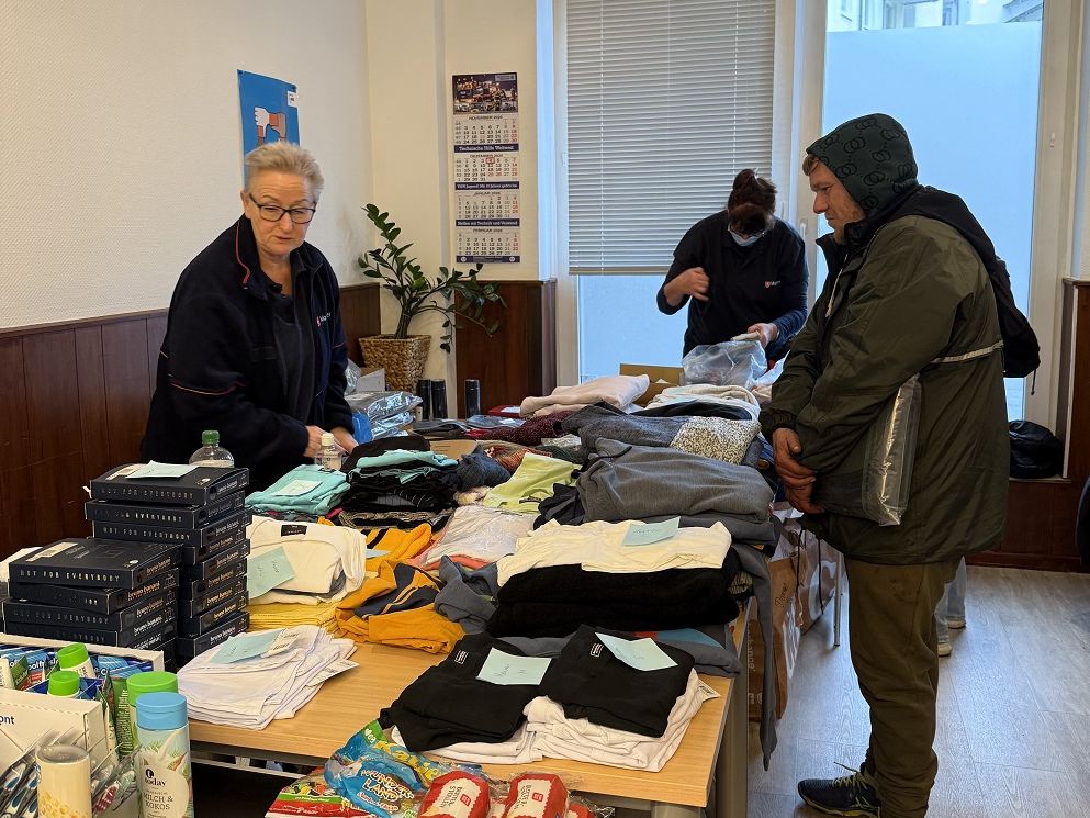 Ein Frau in Malteser Kleidung steht hinter Tische, auf denen verschiedene Stapel warmer Kleidung liegen. Vor dem Tisch steht ein Mann in Kapuzenjacke.