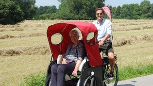 Ein Mann in Lederhose steuert eine Malteser Rikscha mit rotem Dach, vorne sitzt eine ältere Dame, im Hintergrund ein Getreidefeld