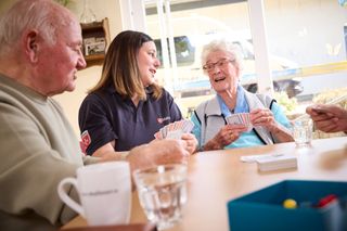 Mehrere Personen sitzen gemeinsam an einem Tisch und spielen Karten. Auf dem Tisch stehen Gläser und eine Tasse. Im Hintergrund fällt durch ein großes Fenster Tageslicht in den Raum, draußen ist ein Fahrzeug der Malteser zu erkennen.