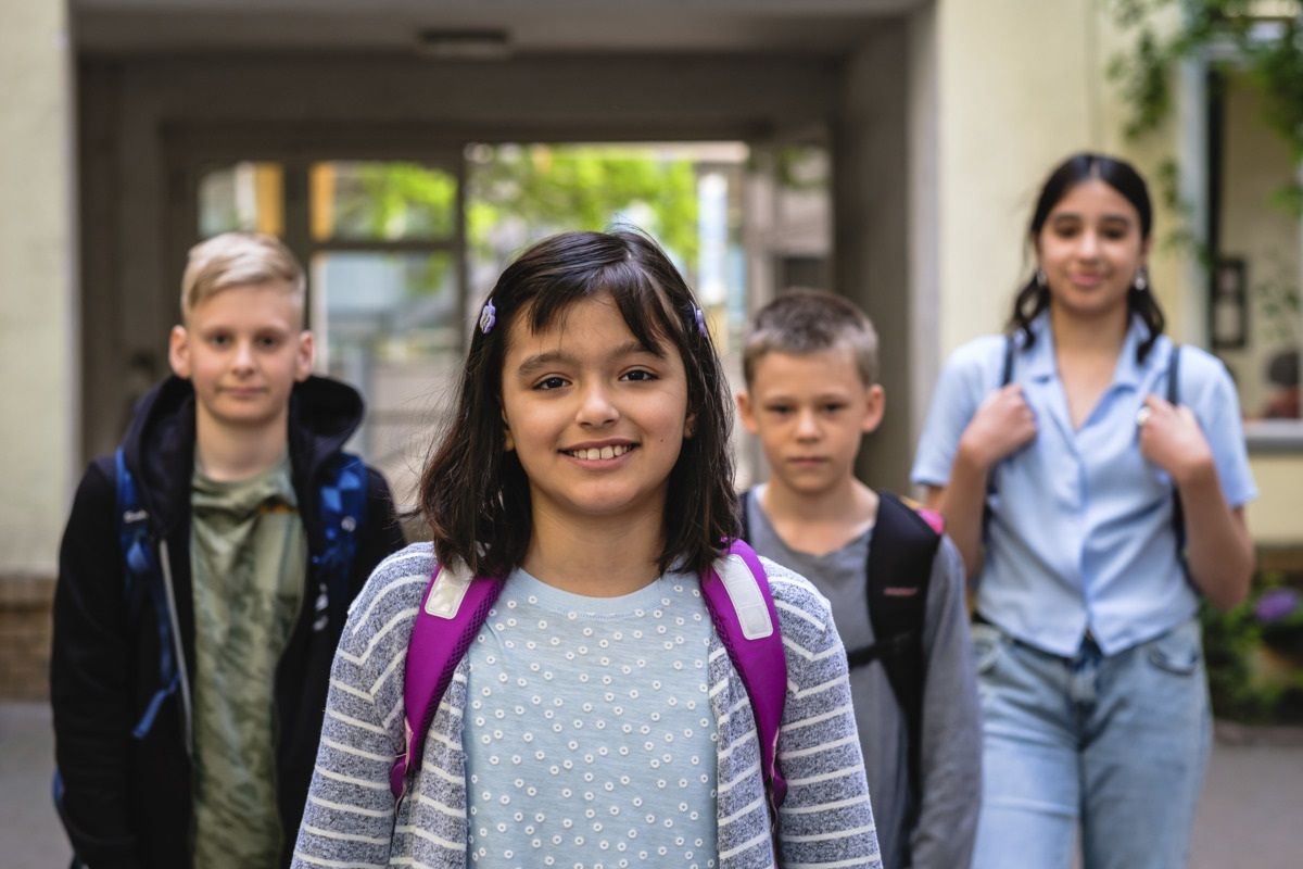 Vier Kinder stehen vor einem Eingang eines Gebäudes. Alle haben einen Rucksack auf. Das Kind im Vordergrund lächelt.