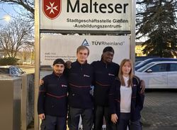 Vier junge Menschen mit Malteser Jacke stehe vor einem Malteser Schild