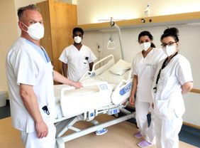 Vier Pflegekräfte in weißer Dienstkleidung und Maske an einem Klinikbett in einem Patientenzimmer.