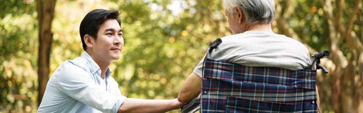 Junger Mann kniet vor einer älteren Person im Rollstuhl