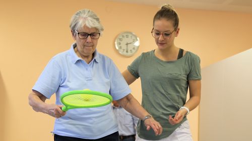 Ältere Frau balanciert einen Ball auf einem Schläger, daneben steht eine betreuende Person zur Unterstützung.
