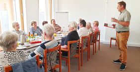 Ein Mann spricht zu mehreren Frauen und Männern, die an einem langen Tisch sitzen.