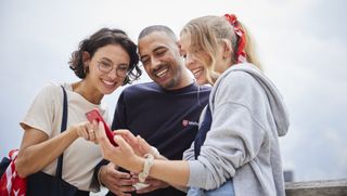 Drei junge Malteser Mitarbeitende, jeglichen Aussehens und Geschlechts, schauen lächelnd auf ein Handy, dass sie gemeinsam mittig vor sich halten.