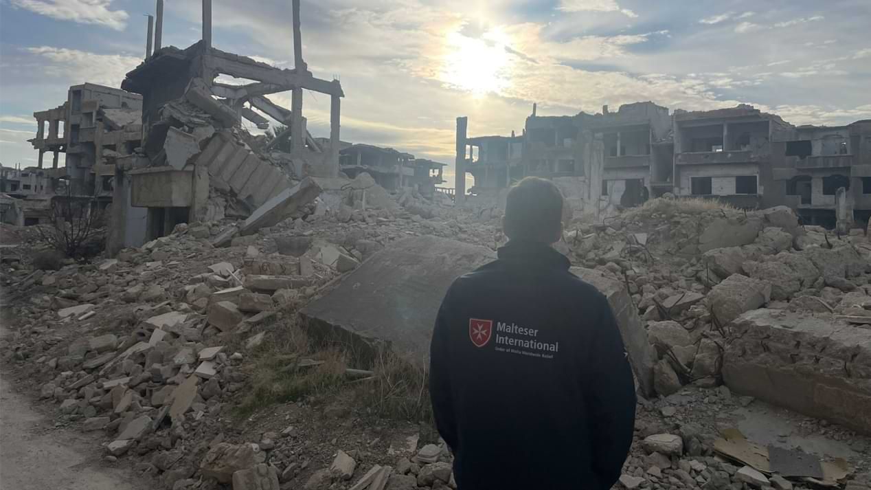 Ein Mann mit einer Jacke von „Malteser International“ steht mit dem Rücken zur Kamera vor einer Gebäuderuine in Syrien.