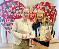 Zwei Frauen stehen vor einem roten Herz, in den Händen halten sie Geschenke