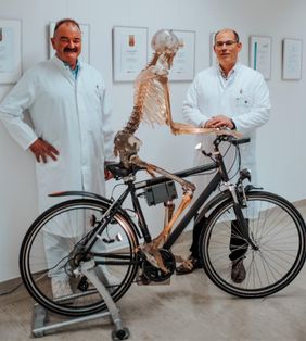 Zwei Ärzte in weißen Kitteln stehen neben einem Skelett auf einem Fahrrad.
