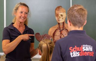 Dozent*in zeigt auf Skelett, während ihr zwei jüngere Menschen zuhören