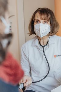 Frau Dr. Anne Rauhut mit einer Maske sitzt mit dem Gesicht frontal zu einem Patienten, den man im Vordergrund erkennen kann. Sie hält ein Stethoskop in der Hand. 