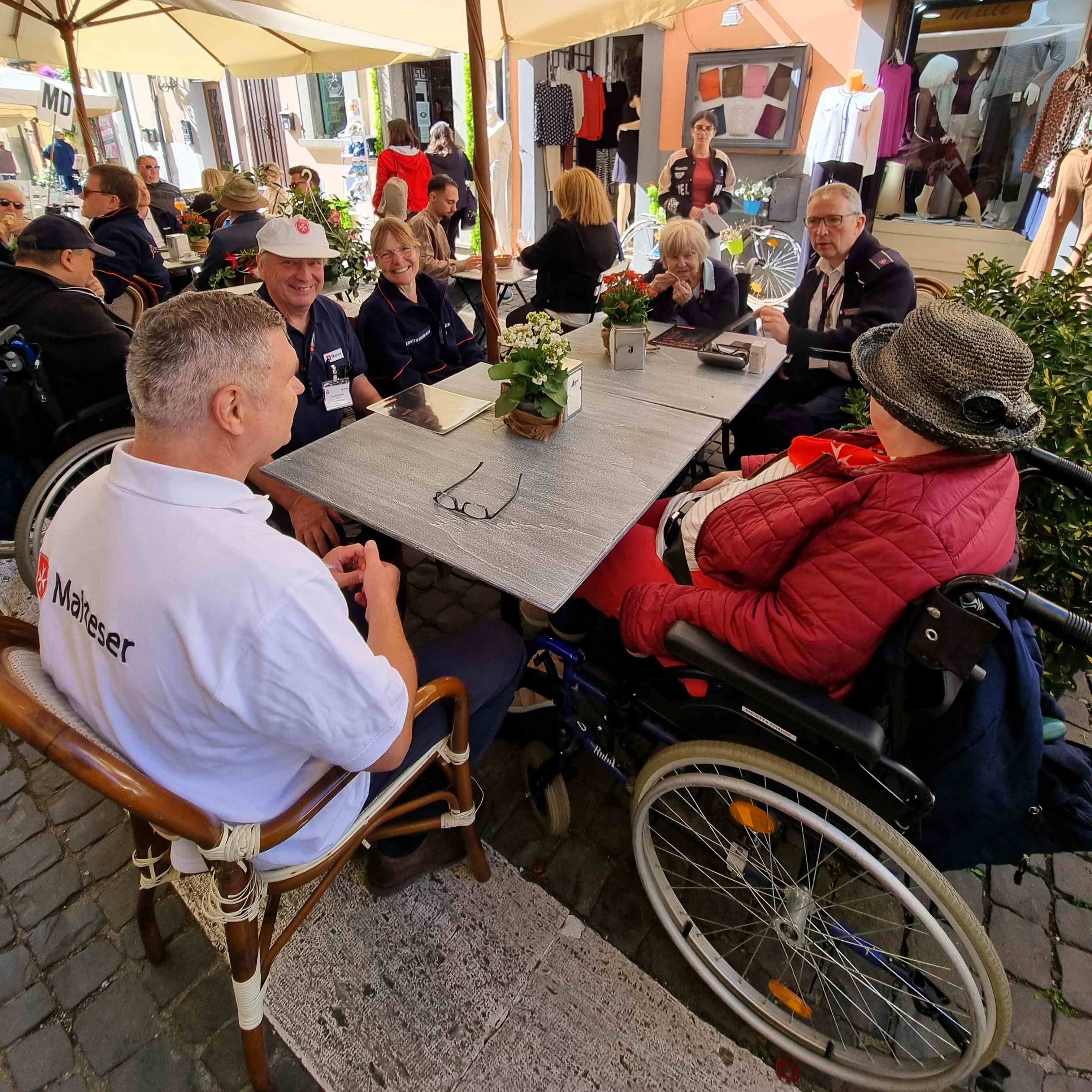 Menschen sitzen an einem Tisch im Freien in Castel Gandolfo, eine Person im Rollstuhl trägt eine rote Jacke und einen Hut.