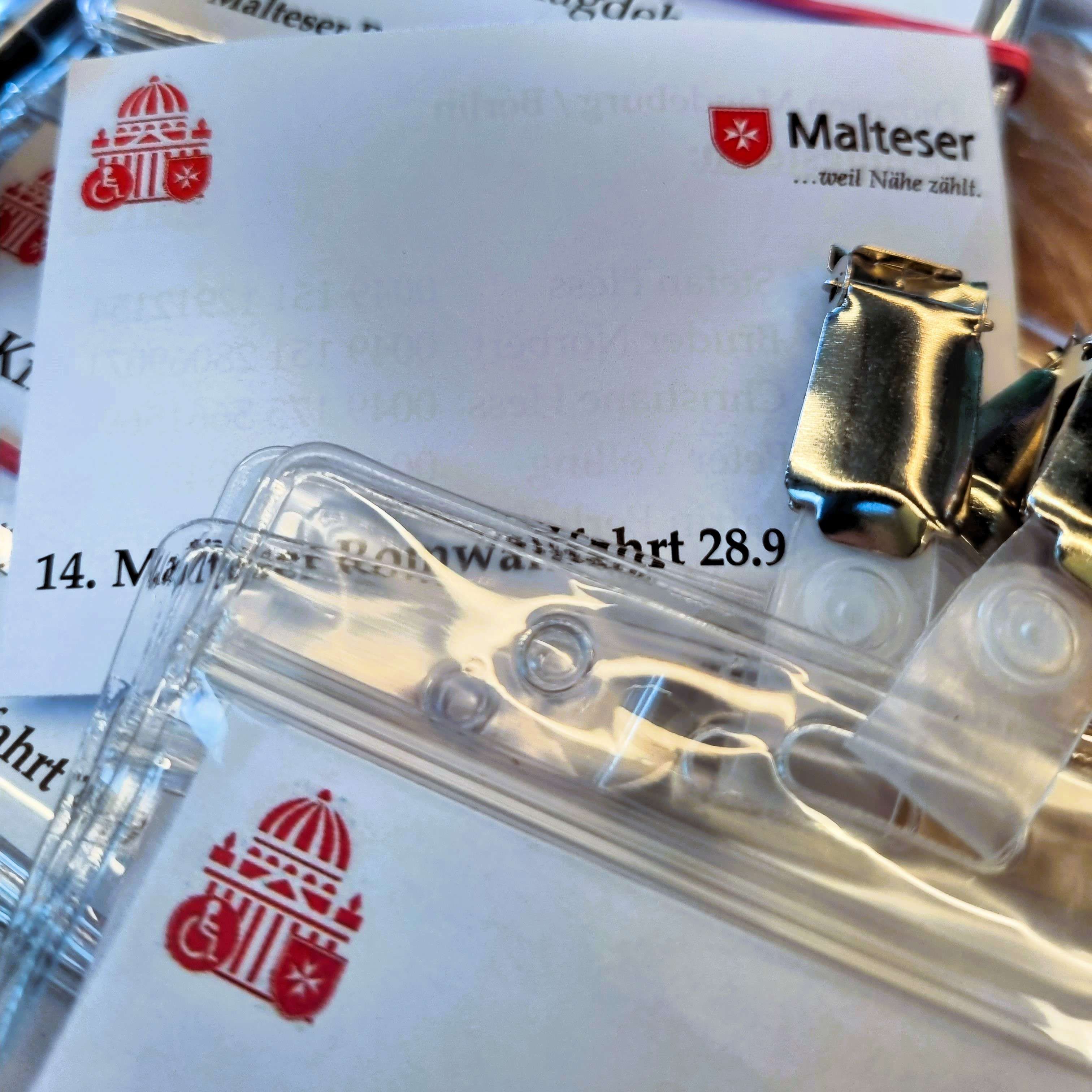 Malteser-Logo auf mehreren weißen Papieren und durchsichtigen Namensschildern mit Metallclip, teilweise überlappend.