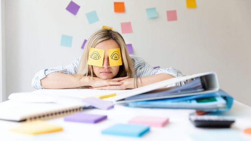 Frau in hellem Büro mit bunten Klebezetteln lehnt gelangweilt auf einem Schreibtisch.