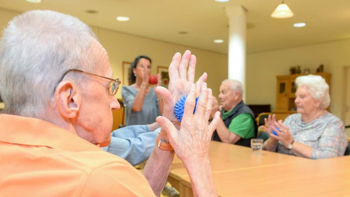 Eine Gruppe älterer Menschen hat Bälle zwischen den Händen und rollen diese hoch und runter