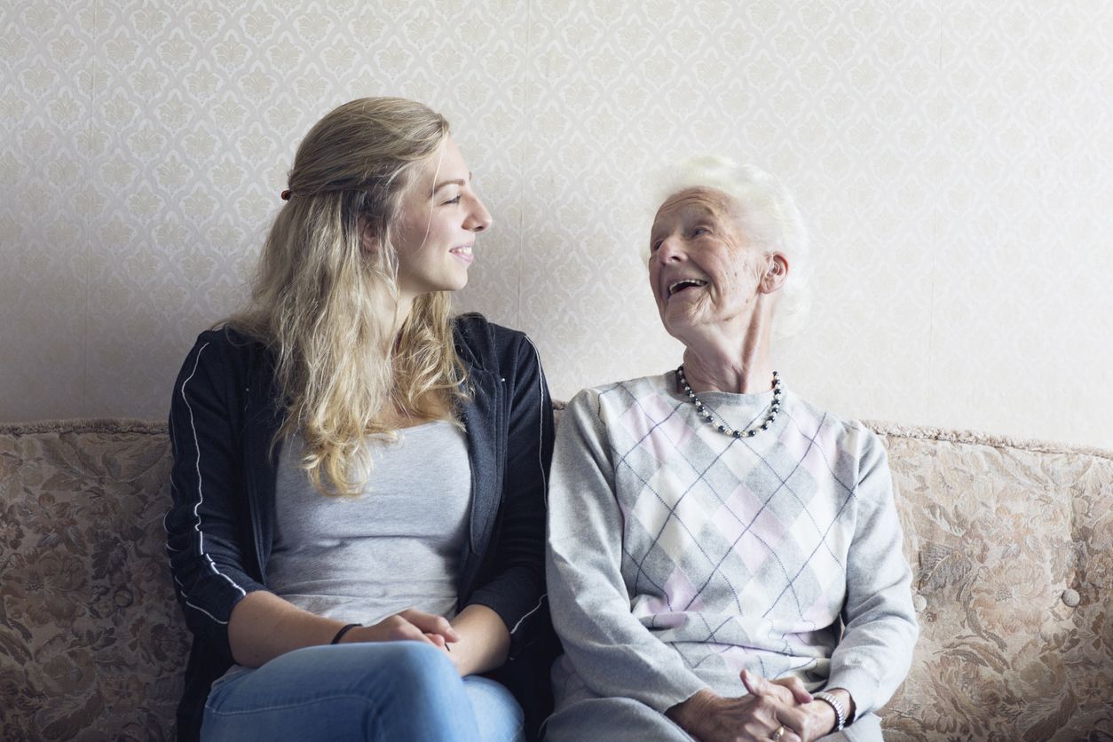 Junge Frau und Seniorin lächeln sich sitzend auf einem Sofa an.