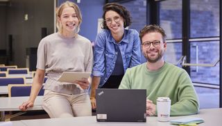 Drei junge Menschen – zwei Frauen und ein Mann – befinden sich in einem Schulungsraum der Malteser. Der Mann sitzt vor einem geöffneten Laptop am Tisch und die beiden Frauen sitzen links hinter ihm auf der Tischplatte. Alle drei lächeln in die Kamer