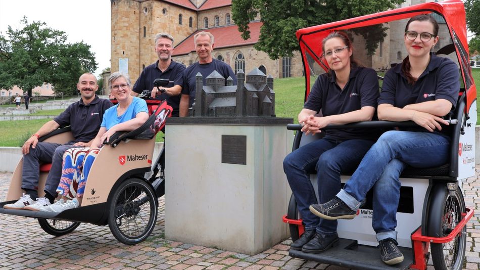 Sechs Personen posieren gemeinsam im Freien vor einer großen Kirche. Zwei von ihnen sitzen in rikschaähnlichen Fahrrädern mit „Malteser“-Aufschrift, während die anderen danebenstehen. In der Mitte steht ein kleines Modell eines Gebäudes auf einem Sockel, umgeben von gepflastertem Boden und Grünflächen.