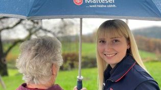 Malteser Mitarbeiterin hält einen Regenschirm über eine Seniorin