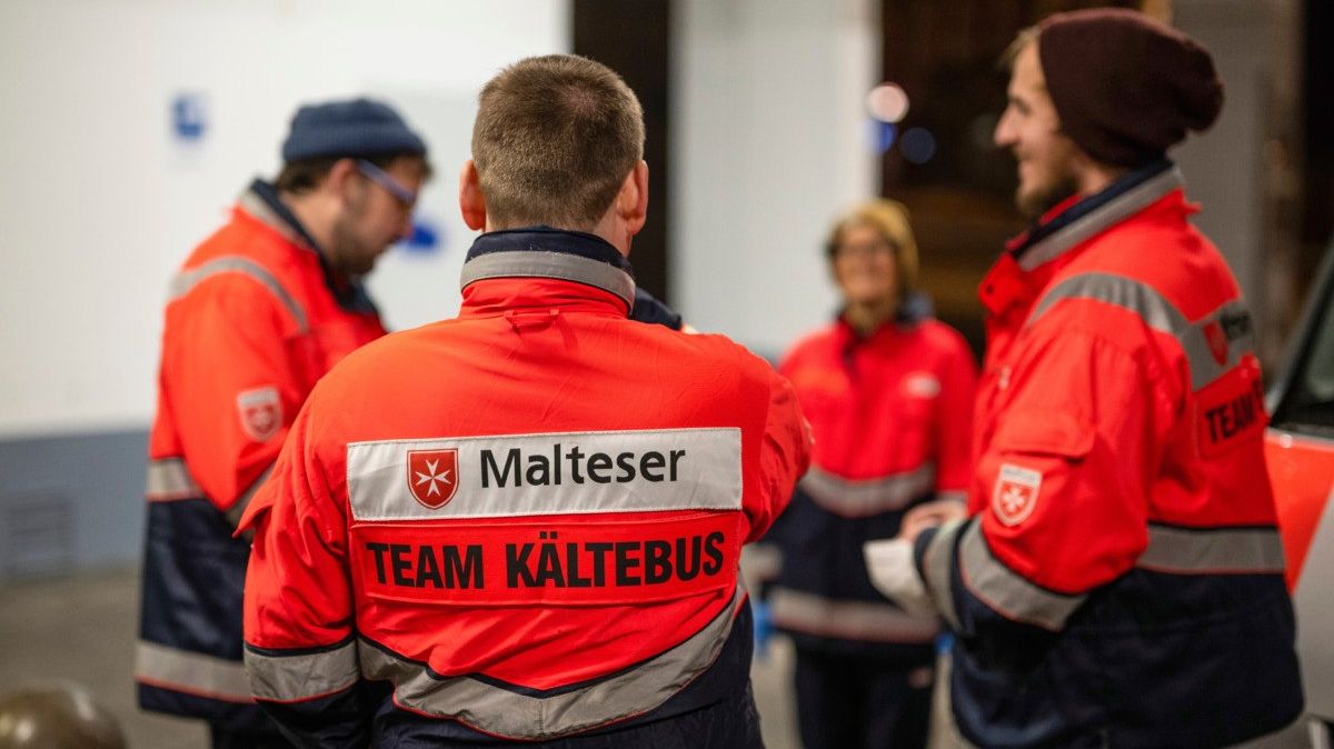 Drei Männer und eine Frau mit Einsatzkleidung der Malteser vor einem Gebäude bei Nacht.