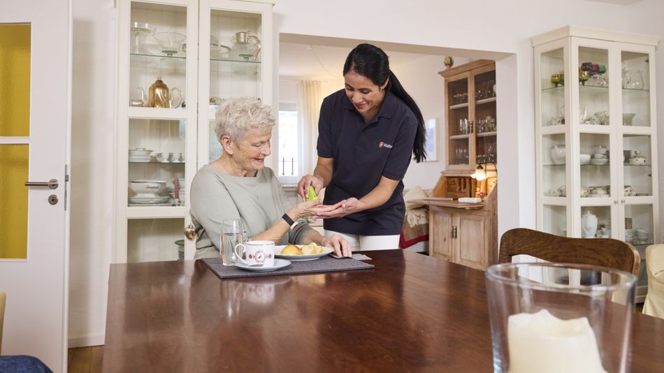 Eine junge, lächelnde Frau hilft einer älteren Frau, die an einem Esstisch sitzt, bei der Dosierung eines Medikaments.