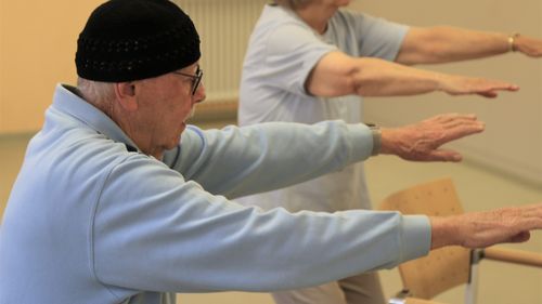 Zwei ältere Personen führen eine Armstreckübung mit nach vorn gestreckten Armen auf Schulterhöhe aus.