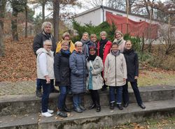 Eine Gruppe von Menschen steht draußen auf mehreren Steinstufen zusammen. Sie tragen überwiegend herbstliche oder winterliche Kleidung wie Jacken und Schals. Im Hintergrund sind ein Gebäude, Bäume mit herabgefallenen Blättern und Sträucher zu sehen. Die Szene wirkt wie ein Gruppenfoto während einer Veranstaltung oder eines Kurses in herbstlicher Umgebung.