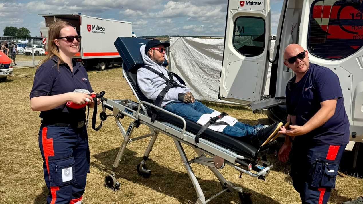 Ein Mann mit Baseballmütze und Sonnenbrille auf einer Rollbahre und zwei junge Menschen in Einsatzkleidung der Malteser auf einem Festivalgelände. 