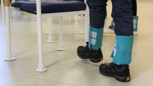 Person mit Gewichtsmanschetten an den Unterschenkeln steht vor einem Stuhl zur Durchführung einer Gleichgewichtsübung.