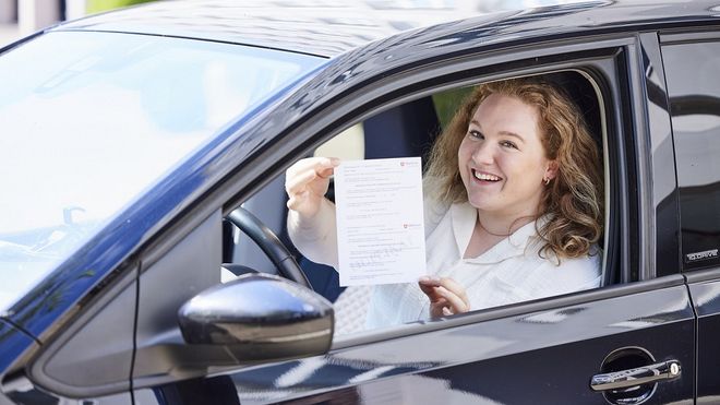 Eine junge Frau sitzt hinter dem Lenkrad eines PKW und hält eine Bescheinigung durch das geöffnete Fenster.