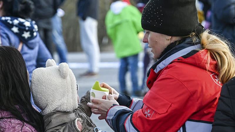 Frau in Malteser Einsatzkleidung gibt einem kleinen Kind auf dem Arm der Mutter etwas zu trinken. 