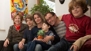 Vater, Mutter und zwei gesunde Kinder sitzen mit einem jüngeren, kranken Kind auf einem Sofa. Neben ihnen die Dame vom Hospizdienst der Malteser